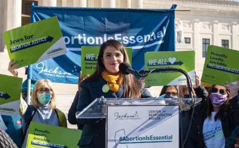 Sheila Katz stands at a podium in front of the U.S. Supreme Court addressing a crowd of protesters. More protesters stand behind her holding signs that read, "Jews for #Abortion Justice."