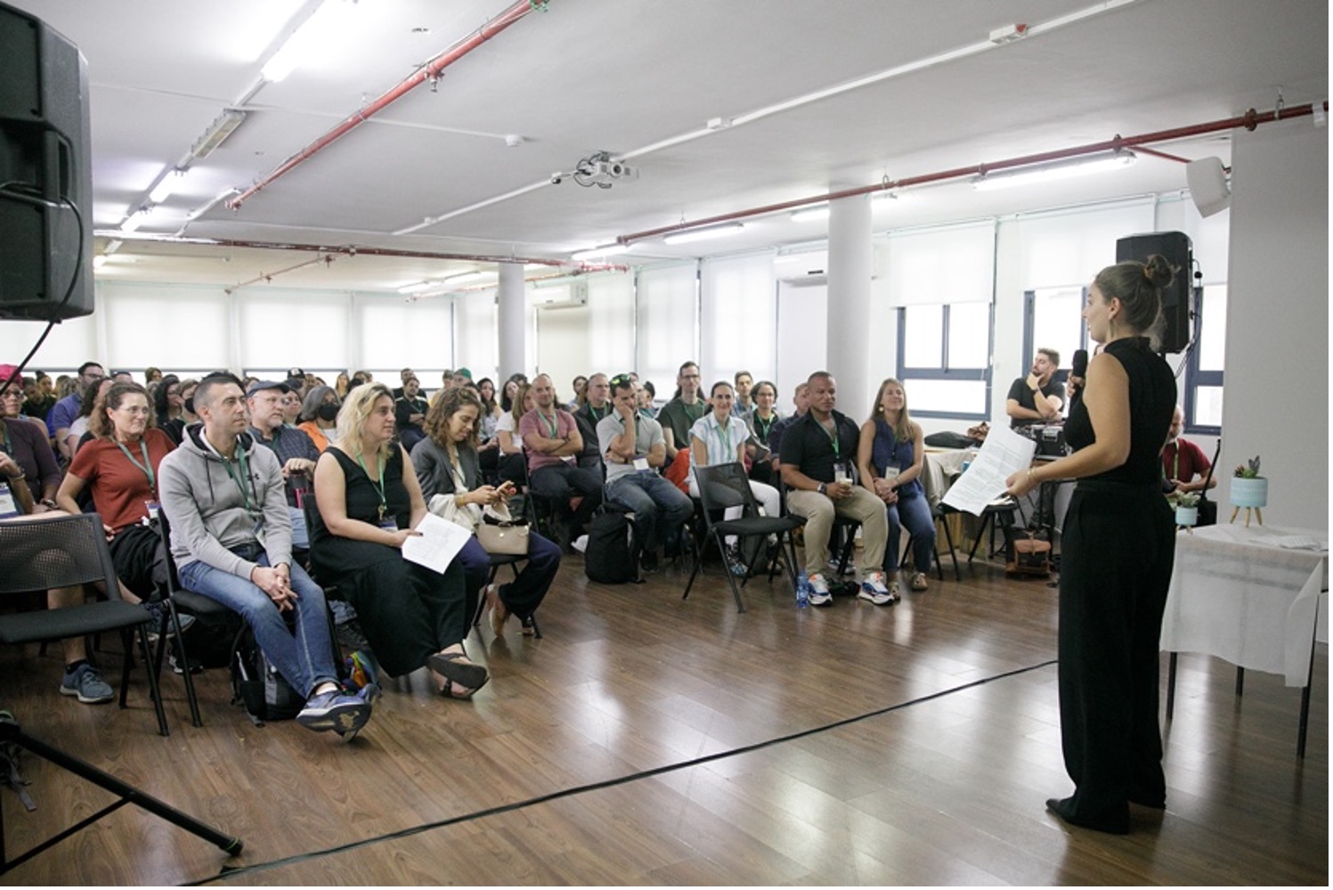 A woman with her hair in a bun stands at the front of a room with a microphone in front of approximately 100 seated adult participants.