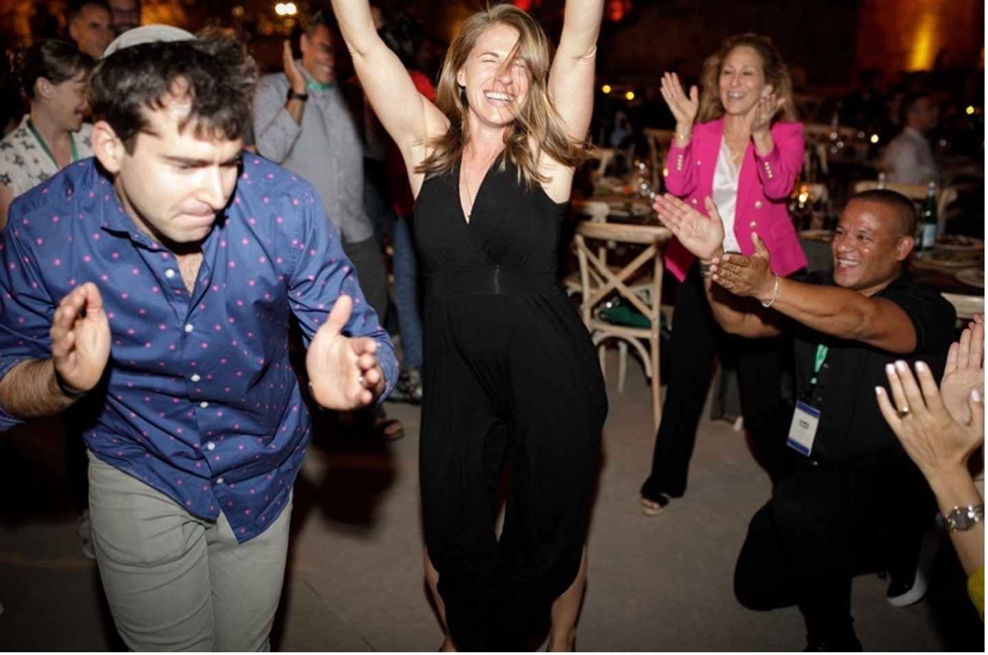 A group of professionals smiling and dancing. A woman in the middle lifts her arms over her head in joy while her colleagues clap for her.