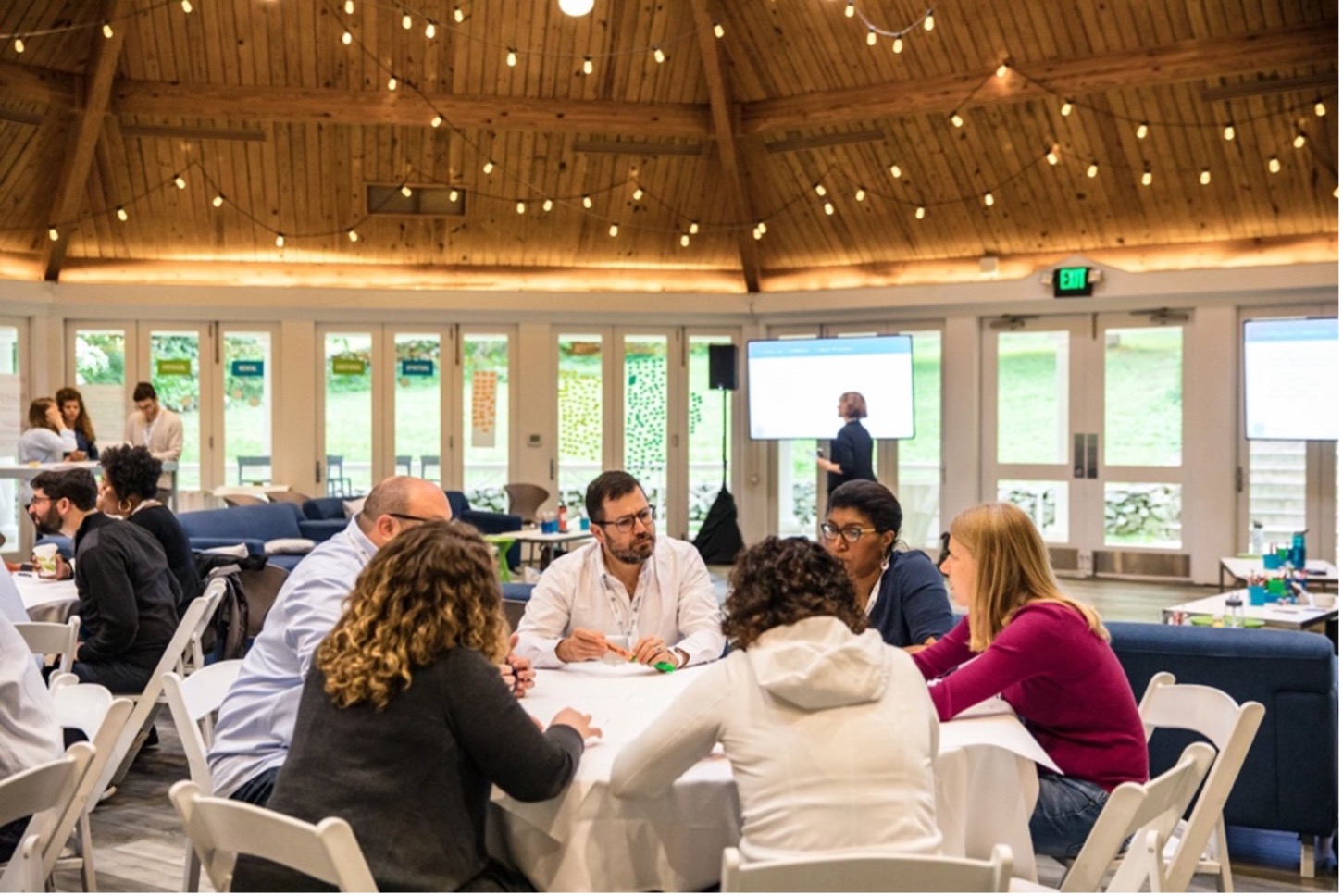 A group of adult program partiicpants sit around a table in deep discussion. Other participants sit around them engaged in their own discussions. The room has high ceilings with twinkle lights strung across the top.